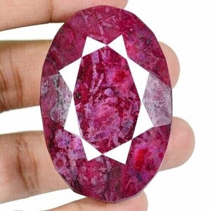 May include: Oval-shaped, faceted ruby gemstone with a deep red hue and white geometric design. The gemstone is held in a hand, showcasing its polished surface and natural inclusions. A close-up view highlights the gem's color and cut.