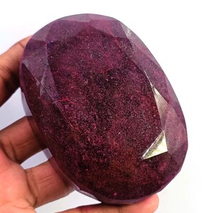 Pode incluir: Uma grande pedra preciosa de rubi facetada com um tom vermelho profundo. A pedra em forma oval tem múltiplas facetas que refletem a luz, criando um efeito brilhante. A pedra preciosa é segurada em uma mão contra um fundo branco.