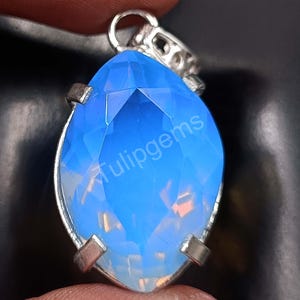 May include: A marquise-shaped pendant with a faceted, light blue gemstone. The gemstone is set in a silver-colored metal frame with a loop at the top for a chain. The text "Tulipgems" is visible on the gemstone.