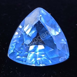 Puede incluir: Una gema triangular facetada de color azul claro. La superficie pulida tiene múltiples facetas que reflejan la luz, creando un efecto brillante. La gema está sobre un fondo oscuro. La forma es triangular.