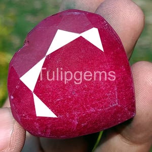 Pode incluir: Uma pedra preciosa vermelha escura facetada em forma de coração com formas geométricas brancas. O texto "Tulipgems" está impresso em branco no centro da pedra. A pedra preciosa é segurada em uma mão.