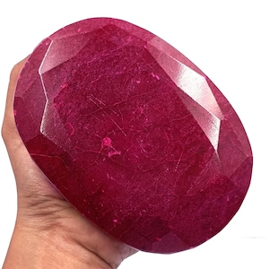 Puede incluir: Gran gema de rubí facetada. La gema de color rojo intenso tiene una superficie pulida con múltiples facetas, creando una brillante exhibición de luz. El rubí se sostiene en una mano, mostrando su tamaño y color.