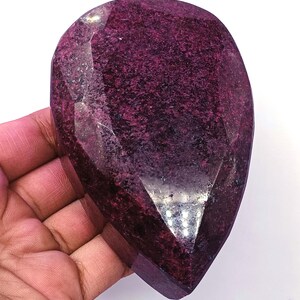 Puede incluir: Una gema facetada en forma de lágrima, de color burdeos intenso. La gema tiene una superficie pulida y reflectante con texturas internas y facetas visibles. La piedra se sostiene en una mano sobre un fondo blanco.
