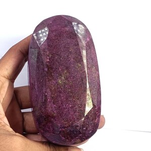 May include: A large, oval-shaped, faceted gemstone with a deep magenta and dark purple hue. The stone features a polished, multi-faceted surface that reflects light, showcasing a textured, crystalline interior with subtle variations in color.