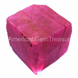 Pode incluir: Uma gema vibrante, de cor fúcsia, cortada em forma de cubo. A superfície tem uma textura áspera e natural com grãos visíveis. O texto "American Gem Treasure" está impresso na lateral. Este item é provavelmente uma gema bruta, não cortada.