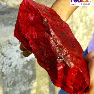 Puede incluir: Una gran gema roja oscura, toscamente tallada, se sostiene en las manos de una persona. La piedra tiene una superficie texturizada y una forma irregular. El logotipo de FedEx Express es visible en la esquina superior derecha.
