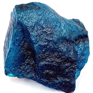 May include: A large, rough-hewn, blue-colored gemstone with a textured surface. The stone has varying shades of blue, from a deep, dark blue to a lighter, brighter blue. The gemstone is displayed against a white background.