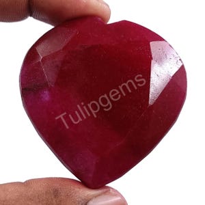 May include: A heart-shaped, faceted, deep red gemstone. The gem has a polished surface with multiple facets, catching the light. The text "Tulipgems" is printed on the surface of the gemstone.
