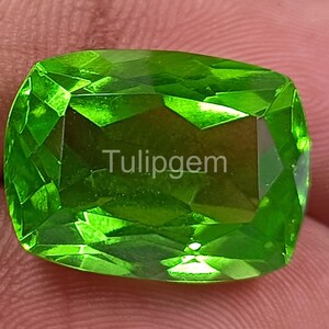 Puede incluir: Una gema verde vibrante y facetada con el texto "Tulipgem" visible. La gema tiene un corte cojín rectangular y refleja la luz. El color es verde lima brillante. La gema está sujeta en la mano de una persona.