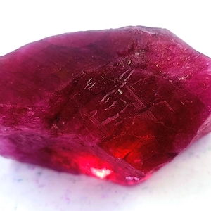 May include: A raw, uncut ruby gemstone with a deep red-pink hue. The natural stone has a rough, textured surface and a slightly translucent appearance, with areas of intense color and light reflection.
