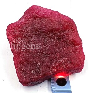 Könnte beinhalten: Ein großer, ungeschliffener Rubin-Edelstein in tiefroter Farbe. Die Oberfläche hat ein strukturiertes Aussehen. Das Wort "lipgems" ist auf der Oberfläche sichtbar. Der Edelstein wird von einem Licht beleuchtet.