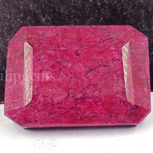 May include: A rectangular, faceted gemstone with a deep magenta hue. The stone has a polished surface with internal veining, and the edges are cut with a geometric design. The gemstone is set against a dark background.