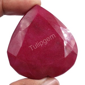 Puede incluir: Una gema de rubí facetada en forma de corazón. La gema es de un color rojo intenso, con múltiples facetas que capturan la luz. El texto "Tulipgem" está impreso en la superficie de la gema.