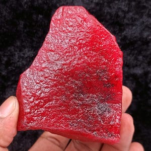 May include: A rough-cut, vibrant red gemstone with a textured surface. The ruby-like stone is held in a hand, showcasing its natural, irregular shape and deep color. The gemstone's surface has a slightly reflective quality.