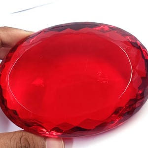 Op de afbeelding: Een grote, ovaalvormige, doorschijnende rode glazen presse-papier met een gefacetteerde rand. Het object heeft een glad, plat middengedeelte omringd door een reeks geometrische, diamantgeslepen facetten die het licht reflecteren.