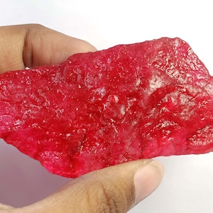 May include: A rough, uncut ruby gemstone with a deep red hue. The raw mineral has a textured surface and irregular shape, held in a hand against a white background. The ruby appears to be a natural, unpolished specimen.