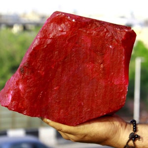 Puede incluir: Una gran gema de rubí en bruto, de color rojo intenso. La piedra se sostiene en una mano, mostrando su tamaño y textura. El fondo está borroso, con indicios de vegetación y edificios.