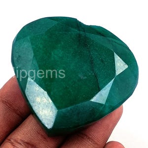 Puede incluir: Una piedra preciosa verde esmeralda en forma de corazón y facetada. La gema tiene una superficie pulida con múltiples facetas, que reflejan la luz. El texto "ipgems" es visible en la superficie de la gema.