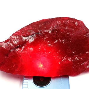 May include: A large, rough-cut ruby gemstone in a deep red hue. The stone has a textured surface and is lit from below, producing a bright glow. The ruby is presented against a white backdrop.