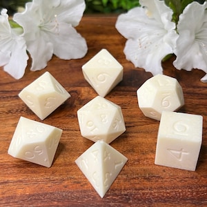 May include: A set of seven white dice with black numbers on a wooden surface. The dice are in the shape of a cube, a pyramid, a tetrahedron, and a dodecahedron.