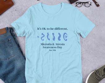 May include: Light blue t-shirt with the text "It's OK to be different." and a graphic representing an ear. The shirt also includes the text "Microtia & Atresia Awareness Day Nov 9th."