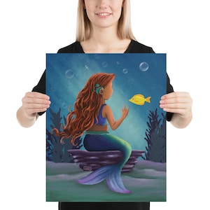Puede incluir: Una sirena con el pelo largo y rojo se sienta en una roca en el océano. Lleva una cola azul y verde y un top azul. Está mirando a un pez amarillo que nada frente a ella.
