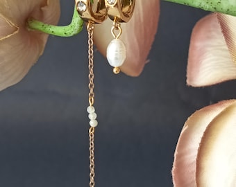 Pode incluir: Brincos de argola dourados com uma corrente pendurada e um pingente de pérola. As argolas têm uma pequena pedra preciosa transparente. A corrente tem pequenas contas brancas redondas. Os brincos são exibidos em uma haste verde com flores rosa e brancas ao fundo.