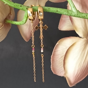 Puede incluir: Pendientes de aro dorados con un colgante de cruz. Cada pendiente presenta una pequeña gema transparente y una cadena con cuentas de colores. Los pendientes se exhiben sobre un fondo oscuro con una flor y un tallo.