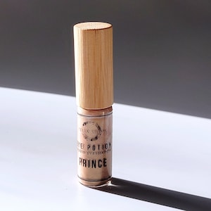 May include: A small glass bottle with a bamboo lid containing a liquid eyeshadow. The bottle has a label that reads "Eye Potion Liquid Eyeshadow Prince" with a circular logo that says "Terra Stoma".