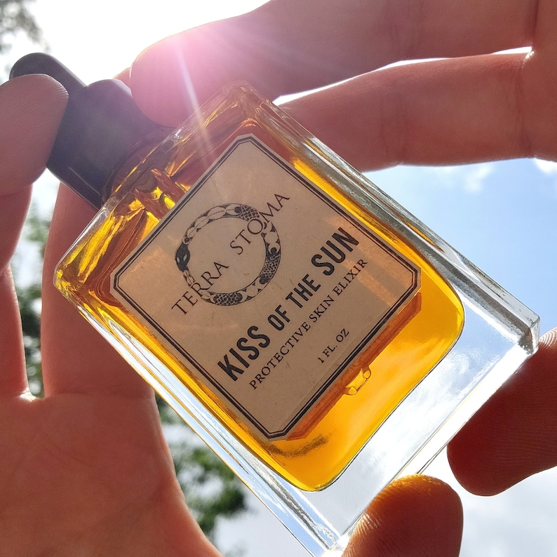 May include: A clear glass bottle with a gold label that reads "Terra Stoma Kiss of the Sun Protective Skin Elixir 1 fl oz". The bottle is filled with a golden liquid and is being held in a hand against a bright blue sky.