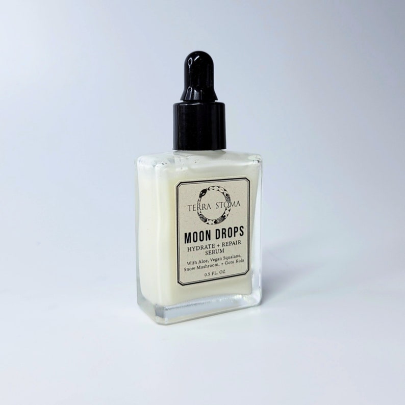 Op de afbeelding: Een doorzichtige glazen fles met een zwarte druppelaar die een witte vloeistof bevat. De fles is gelabeld met "TERRA STOMA MOON DROPS HYDRATE + REPAIR SERUM With Aloe, Vegan Squalane, Snow Mushroom, Gotu Kola 0.5 FL. OZ"
