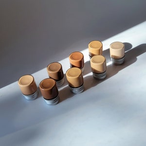 May include: Ten small glass jars with metal lids, filled with different shades of foundation makeup. The jars are arranged in a row on a white surface.