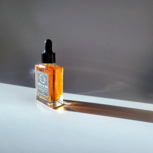 May include: A clear glass bottle with a black dropper top filled with a golden liquid. The bottle is labeled "Terra Firma" and "Kiss of the Sun".