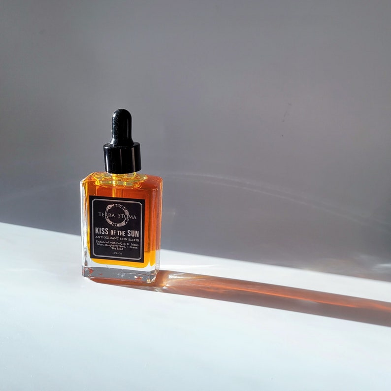 May include: A glass bottle of amber-colored serum with a black dropper top. The bottle is labeled "Kiss of the Sun Antioxidant Skin Elixir" with the brand name "Terra Stigma".