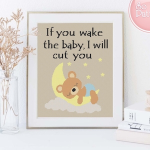 May include: A framed cross-stitch design with a brown bear sleeping on a yellow crescent moon. The text "If you wake the baby, I will cut you" is written in black above the bear.