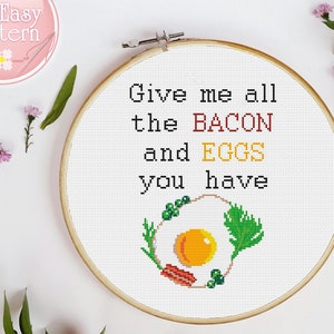Puede incluir: Un aro de bordado blanco con un diseño de punto de cruz que dice "Give me all the BACON and EGGS you have". El diseño incluye un huevo amarillo con una yema roja, tiras de tocino y perejil verde.