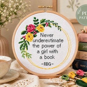 Pu&ograve; includere: Un telaio da ricamo circolare in legno contenente un tessuto bianco con un disegno a punto croce. Il motivo presenta una ghirlanda floreale rosa, gialla e verde che circonda il testo nero: Never underestimate the power of a girl with a book -RBG-.