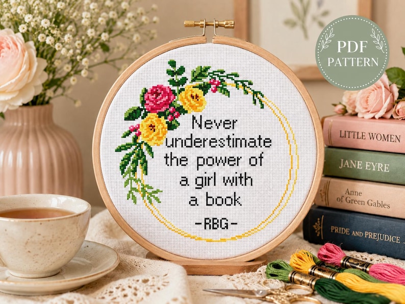 Pu&ograve; includere: Un telaio da ricamo circolare in legno contenente un tessuto bianco con un disegno a punto croce. Il motivo presenta una ghirlanda floreale rosa, gialla e verde che circonda il testo nero: Never underestimate the power of a girl with a book -RBG-.