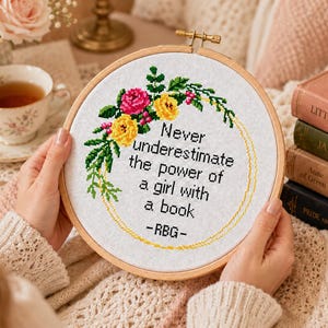 Pu&ograve; includere: Un telaio da ricamo circolare in legno contenente un tessuto bianco con un disegno a punto croce. Il design presenta una corona floreale in rosa e giallo con foglie verdi, che circonda il testo nero: Never underestimate the power of a girl with a book -RBG-.