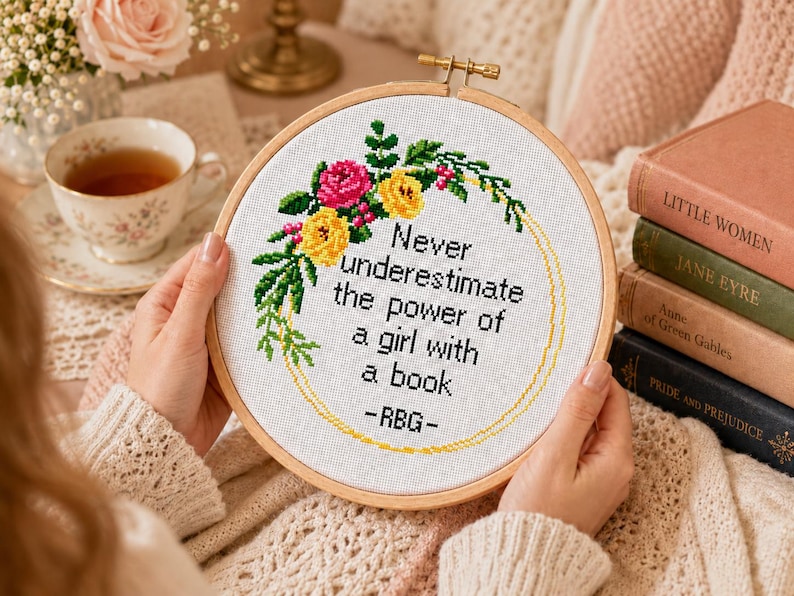 Pu&ograve; includere: Un telaio da ricamo circolare in legno contenente un tessuto bianco con un disegno a punto croce. Il design presenta una corona floreale in rosa e giallo con foglie verdi, che circonda il testo nero: Never underestimate the power of a girl with a book -RBG-.