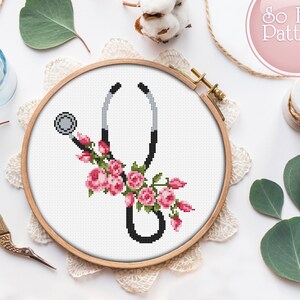 May include: Cross-stitch embroidery in a wooden hoop featuring a stethoscope design adorned with pink roses. The design is set against a white background. The text "So Easy Pattern" is in a pink circle.