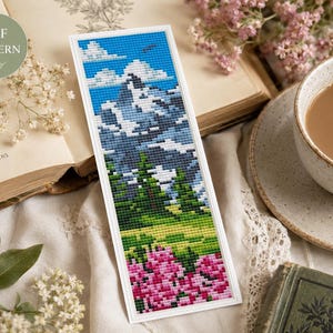 May include: A cross-stitch bookmark featuring a mountain landscape with a blue sky, white clouds, and green trees. The bottom of the bookmark has pink flowers. The bookmark is set on a white background with a cup of coffee and an open book.