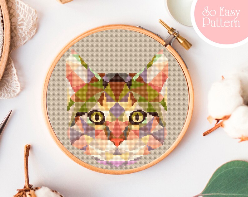 May include: A geometric cross-stitch pattern of a cat's face in a wooden embroidery hoop. The design features a mosaic of triangles in shades of green, orange, pink, and purple. The text "So Easy Pattern" is in the upper right corner.