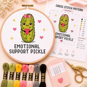 May include: A cross stitch project in a wooden hoop featuring a green pickle with a pink bow holding a red heart, with the text "EMOTIONAL SUPPORT PICKLE". Includes a printed pattern sheet, seven skeins of embroidery floss, needles, and scissors.