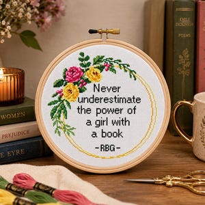 Pu&ograve; includere: Un telaio da ricamo circolare in legno contenente tessuto bianco con un disegno a punto croce. Il design presenta una ghirlanda floreale in rosa, giallo e verde, che circonda il testo nero: Never underestimate the power of a girl with a book -RBG-.