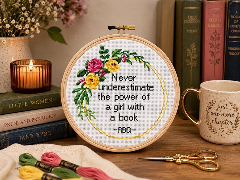 Pu&ograve; includere: Un telaio da ricamo circolare in legno contenente tessuto bianco con un disegno a punto croce. Il design presenta una ghirlanda floreale in rosa, giallo e verde, che circonda il testo nero: Never underestimate the power of a girl with a book -RBG-.