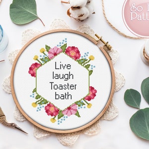 Puede incluir: Un aro de bordado de punto de cruz con un diseño de corona de flores. La corona está hecha de flores rosas y amarillas y hojas verdes. El texto "Live laugh toaster bath" está bordado en el centro del aro.