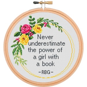 Pu&ograve; includere: Un telaio da ricamo circolare in legno contenente tessuto bianco con un disegno a punto croce. Il testo recita Never underestimate the power of a girl with a book - RBG. Una ghirlanda floreale di rose rosa e gialle incornicia il testo.