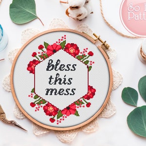 Może przedstawiać: Obręcz do haftu krzyżykowego z białym tłem i czerwoną bordurą kwiatową. Tekst "bless this mess" jest wyszyty czarną nicią w środku obręczy.