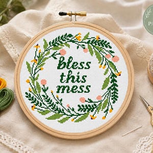 Pode incluir: Um bastidor de bordado circular de madeira contendo um padrão de ponto cruz de uma coroa de folhas verdes com pequenas flores cor-de-rosa e amarelas. O texto "bless this mess" está bordado em letras verde-escuras no centro.